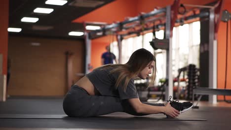 Young-beautiful-yoga-woman-sitting-on-the-floor-on-a-mat-and-stretches-leaning-torso-towards-legs.-Young-girl-doing-yoga-pose-exercise-pilates-stretching.-Modern-gym-with-variety-of-equipment-on-background