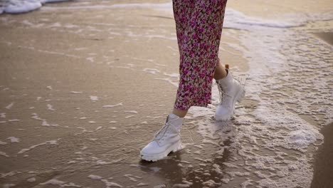 girl in elegant clothes walks along the seashore stepping on foamy waves