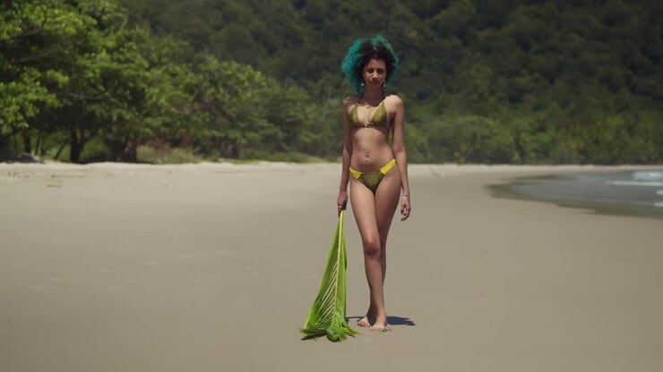 Ragazza in bikini cammina su una spiaggia tropicale con un ramo di palma in mano.