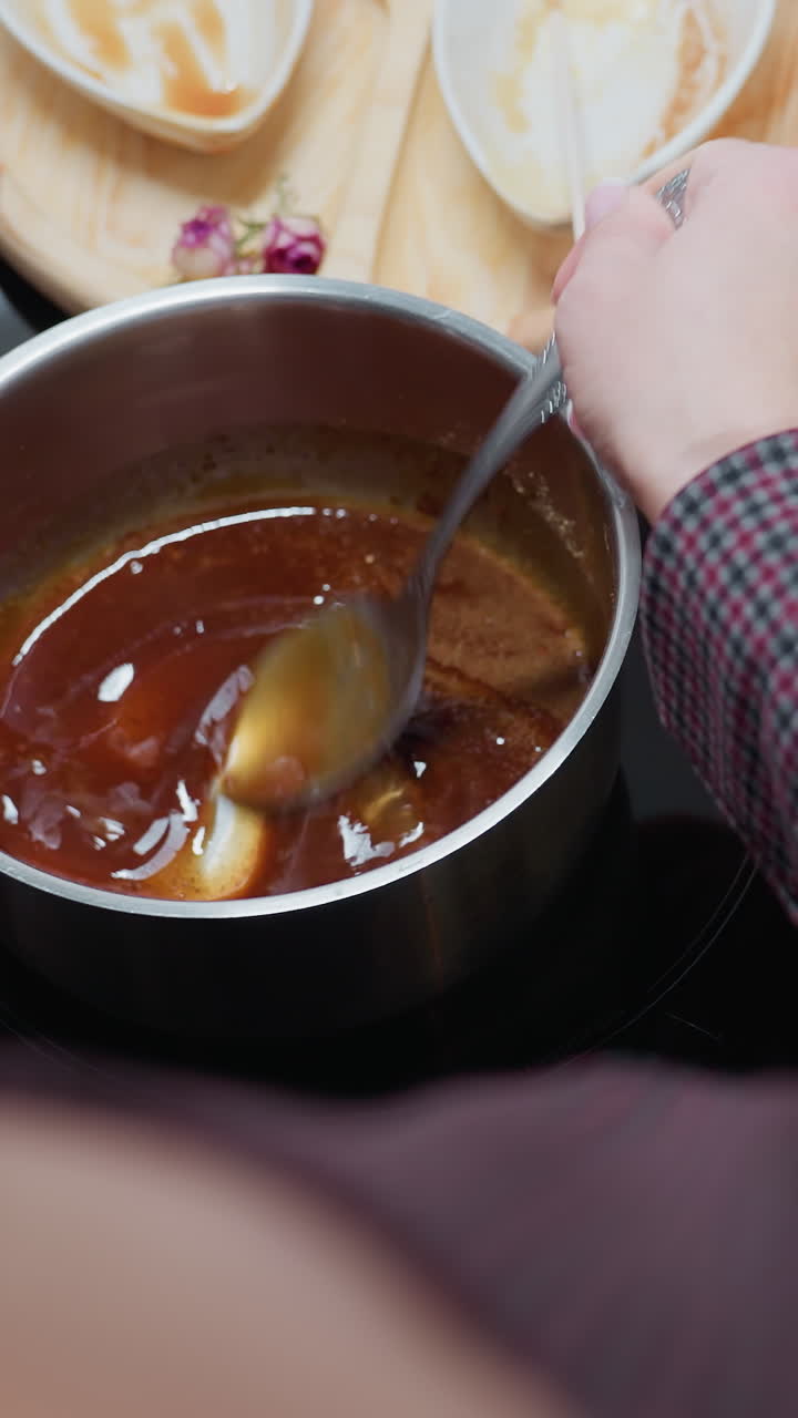 vista trasera de una persona agitando sopa espesa en una olla de acero inoxidable en una estufa de inducción mientras ajusta la temperatura de cocción, con bandeja de madera, cuencos usados y utensilios dispuestos en una encimera de mármol oscuro