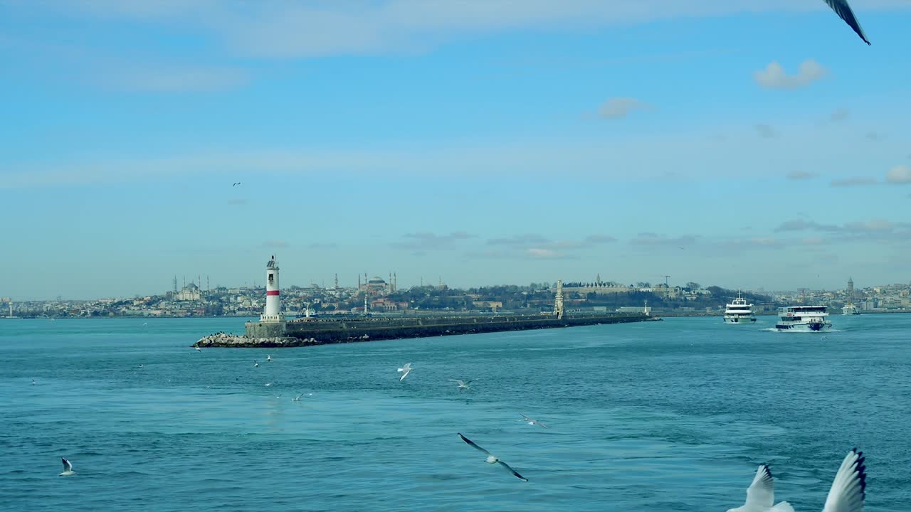 Flight around modern white and red lighthouse in Istanbul harbour with flying seagulls. Mediterranean sea calm waves many concrete buoys pier breakwater navigation shipping port City landscape skyline