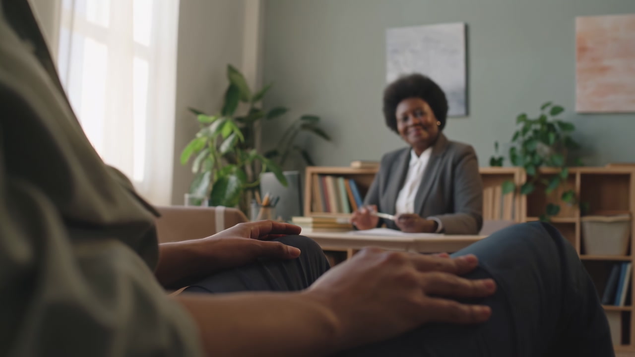 Therapist Consultation Client Hands In Foreground, Counselor Smiles Across Desk Amid Bookshelf And Potted