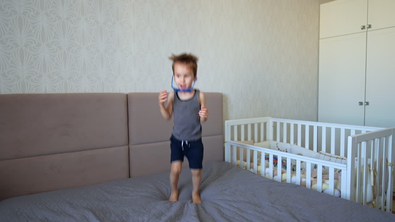 Active toddler boy gets out of his crib on the big double bed. Kid puts on sunglasses and starts to jump happily.