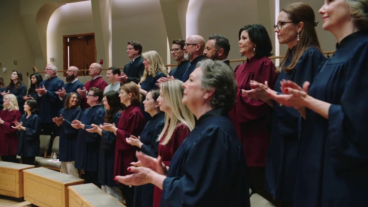 Coro che applaude in risposta alla fine del canto in cappella su gradinate a più livelli, indossando tuniche blu scuro e bordeaux.
