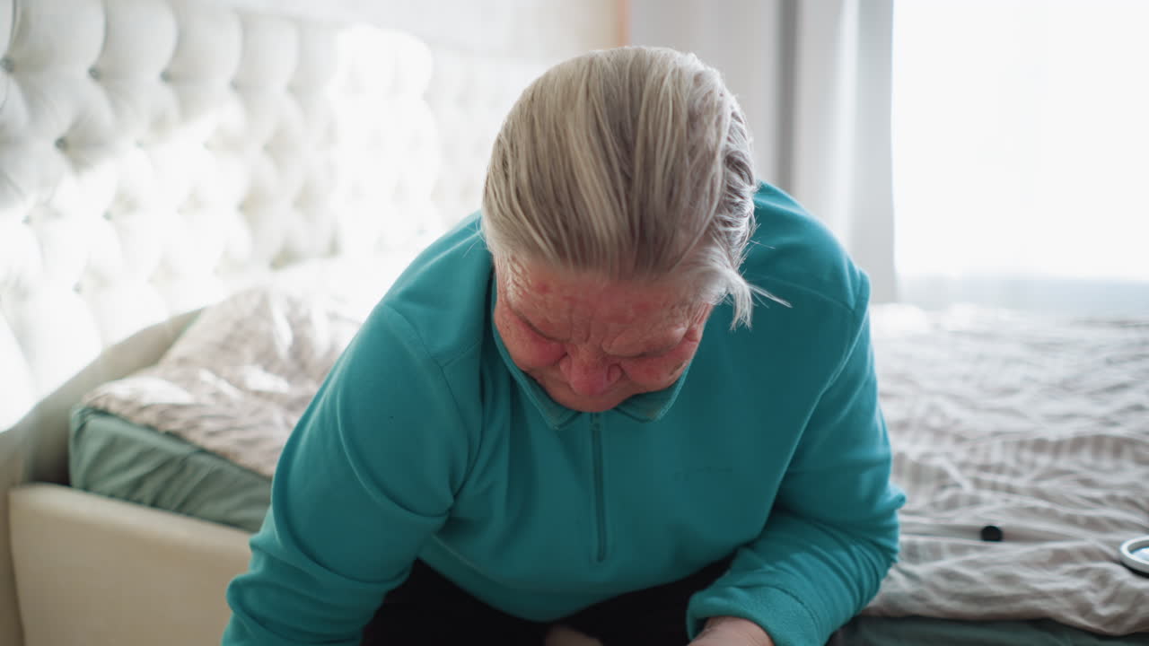 Old woman in green leaning forward, focused on applying ointment to her foot. Soft light from the window illuminates the room, creating a calm, soothing atmosphere for her self-care routine