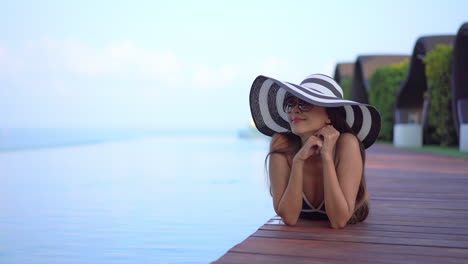 Mujer-Asiática-Sonriente-Con-Un-Gran-Sombrero-A-Rayas-Y-Gafas-De-Sol-Antiguas-Tomando-El-Sol-Junto-A-La-Piscina