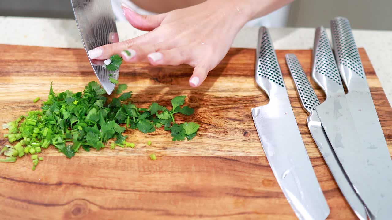 A person skillfully chops fresh coriander on a wooden board with sharp knives, under bright kitchen lighting