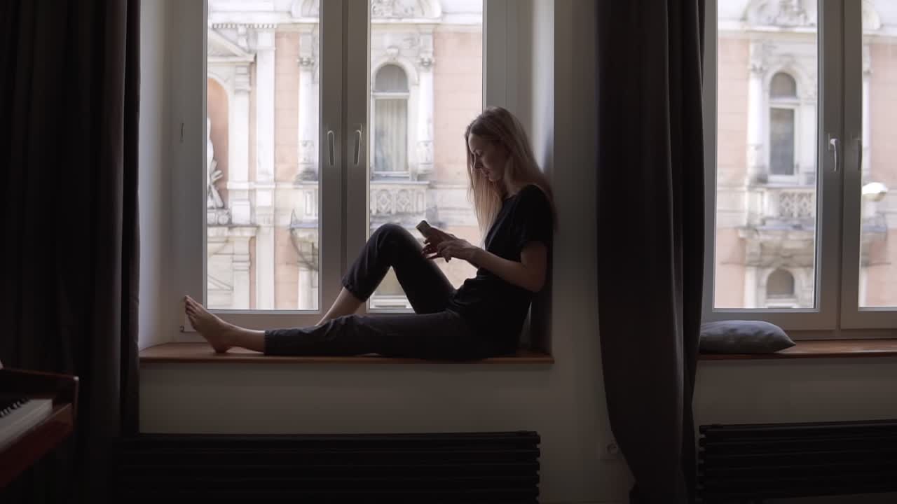 Donna bionda seduta sul davanzale della finestra che usa lo smartphone in soggiorno con pianoforte