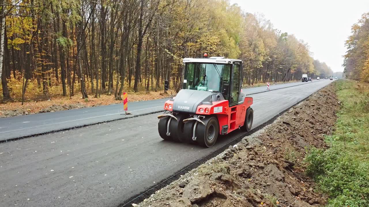 New paver machine at road reconstruction. Asphalt compactor driving on the road in autumn forest. Roadworks in highway outdoors.