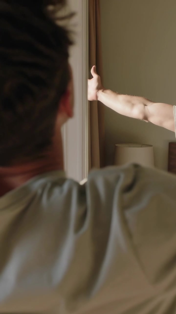 Vídeo vertical: Homem de pé com camiseta cinza observando outro entrar no quarto à direita, dando polegar para cima, lâmpada