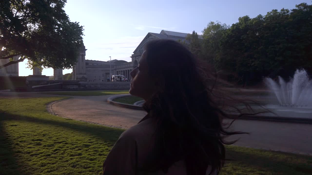 Woman walking near fountain and arch at golden hour. Hair blowing in wind as woman strolls through park