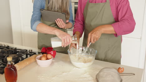 baking, multiracial senior couple mixing ingredients in bowl and using tablet in kitchen, at home