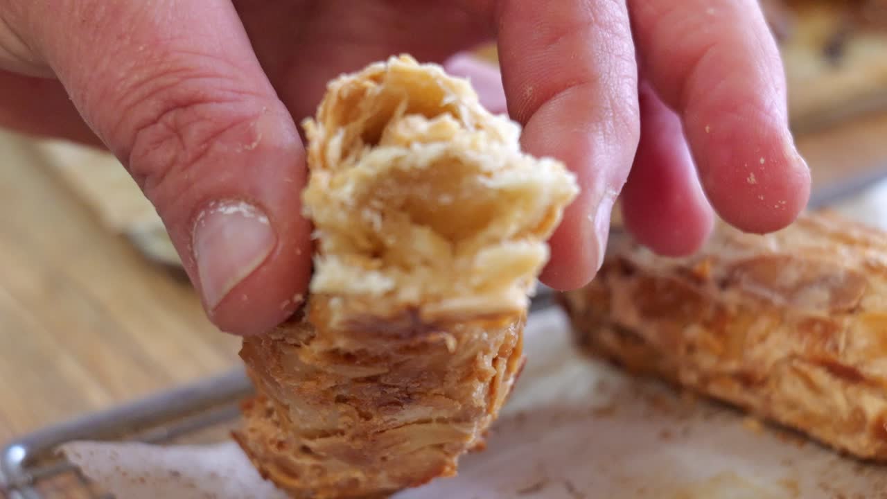 Primo piano di un fornaio che taglia prodotti da forno in pasta sfoglia appena sfornati su una gratella, rivelando l'interno sfogliato in un panificio tradizionale.