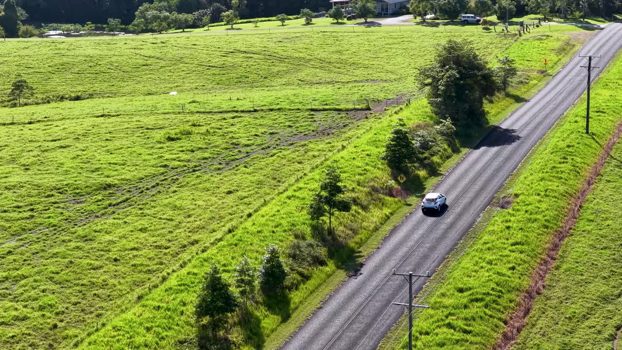 Aerial footage of a car driving through lush green countryside on a sunny day, captured by a drone