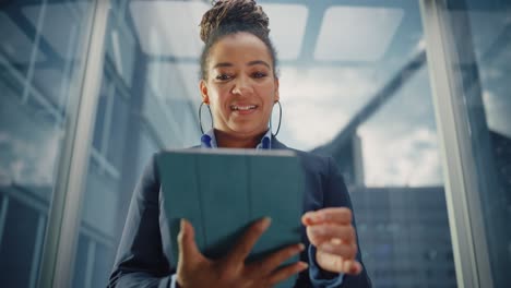 confident black adult female riding glass elevator to office in business center. successful african american manager using tablet computer, write message, check social media and emails in a lift.