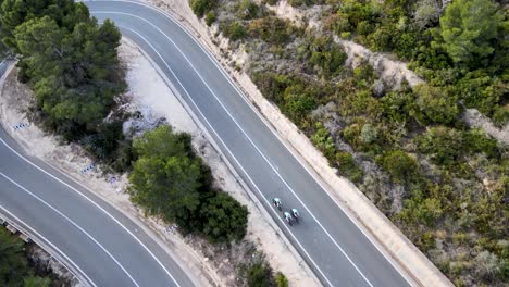 birdsyee drone footage of a team of three cyclists ride up an steep incline on a windy road in calpe, spain