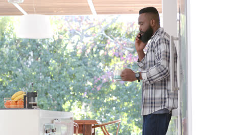 African-american-man-drinking-coffee-and-talking-on-smartphone-in-sunny-kitchen,-slow-motion