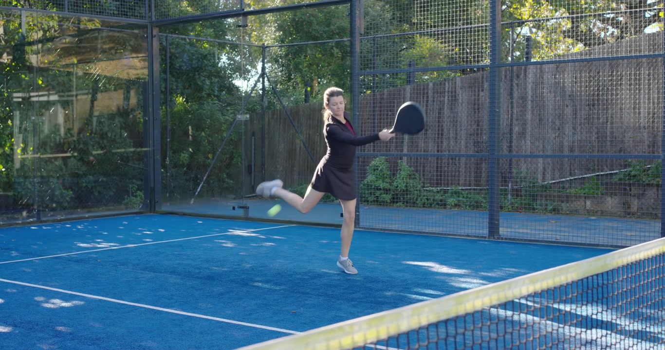 Donna che colpisce di dritto con la racchetta da padel mentre la palla si avvicina su un campo da padel blu, durante un allenamento.