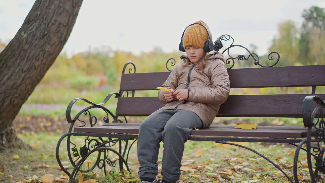 calm kid wearing mustard beanie and over ear headphones sits on ornate iron park bench beneath golden autumn foliage, thoughtfully trims yellow fallen leaf while quietly listening to music