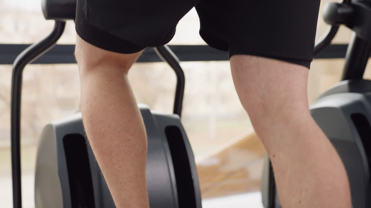 Waist to leg close view of muscular man powering elliptical pedal with strong stride in modern gym next to tall windows during intense cardio session for lower body strength and endurance