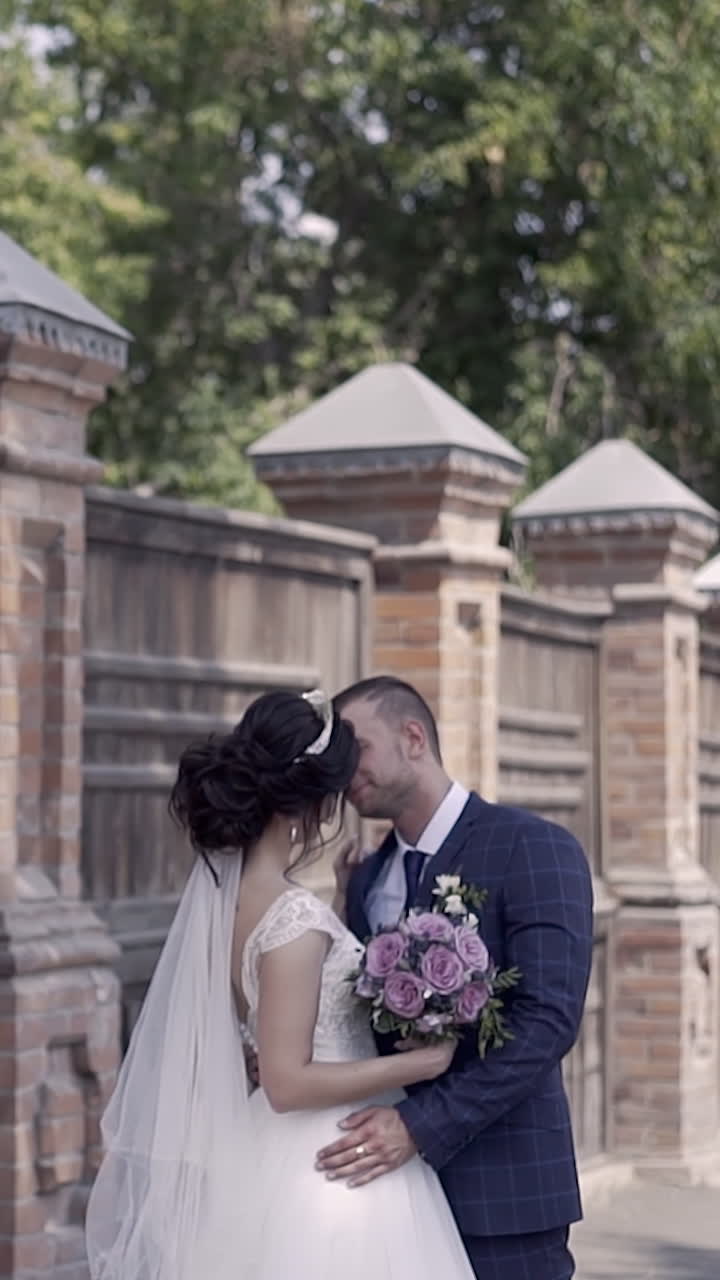 slow motion wonderful wedding couple in celebratory dressing with bouquet hugs against high brown fence