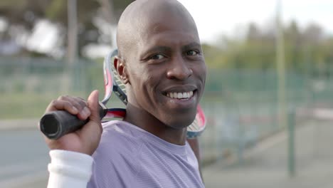 Portrait-of-happy-african-american-male-tennis-player-holding-racket-at-outdoor-court,-slow-motion