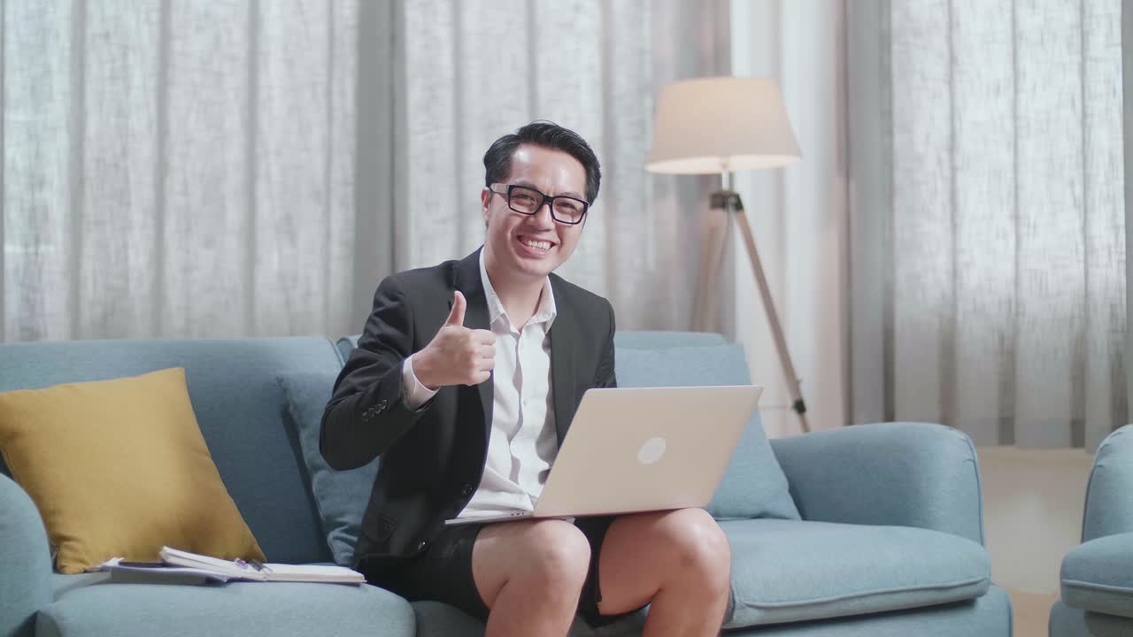 재과 쇼트를 입은 아시아 사업가 가 웃고 집에서 노트북으로 일하면서 엄지손가락을 위로 올려 카메라에 제스처를 하고 있다