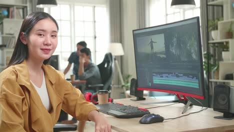 asian female video editor works with footage and sound on his personal computer. she turns and warmly smiles into the camera
