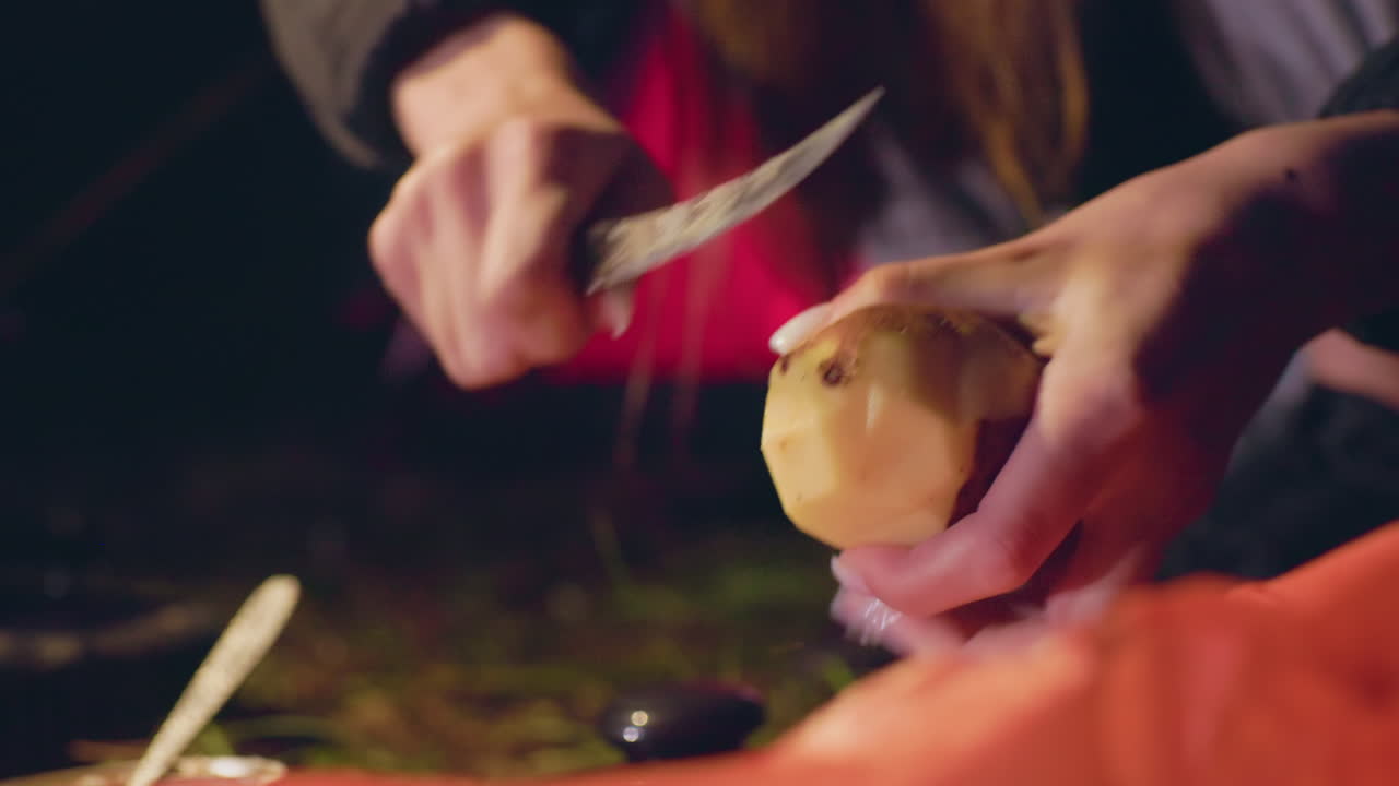 Close up of woman peeling potato with small knife during outdoor night camping, focused hands working under dim light, potato skin curling away as she prepares ingredients for meal near forest tent