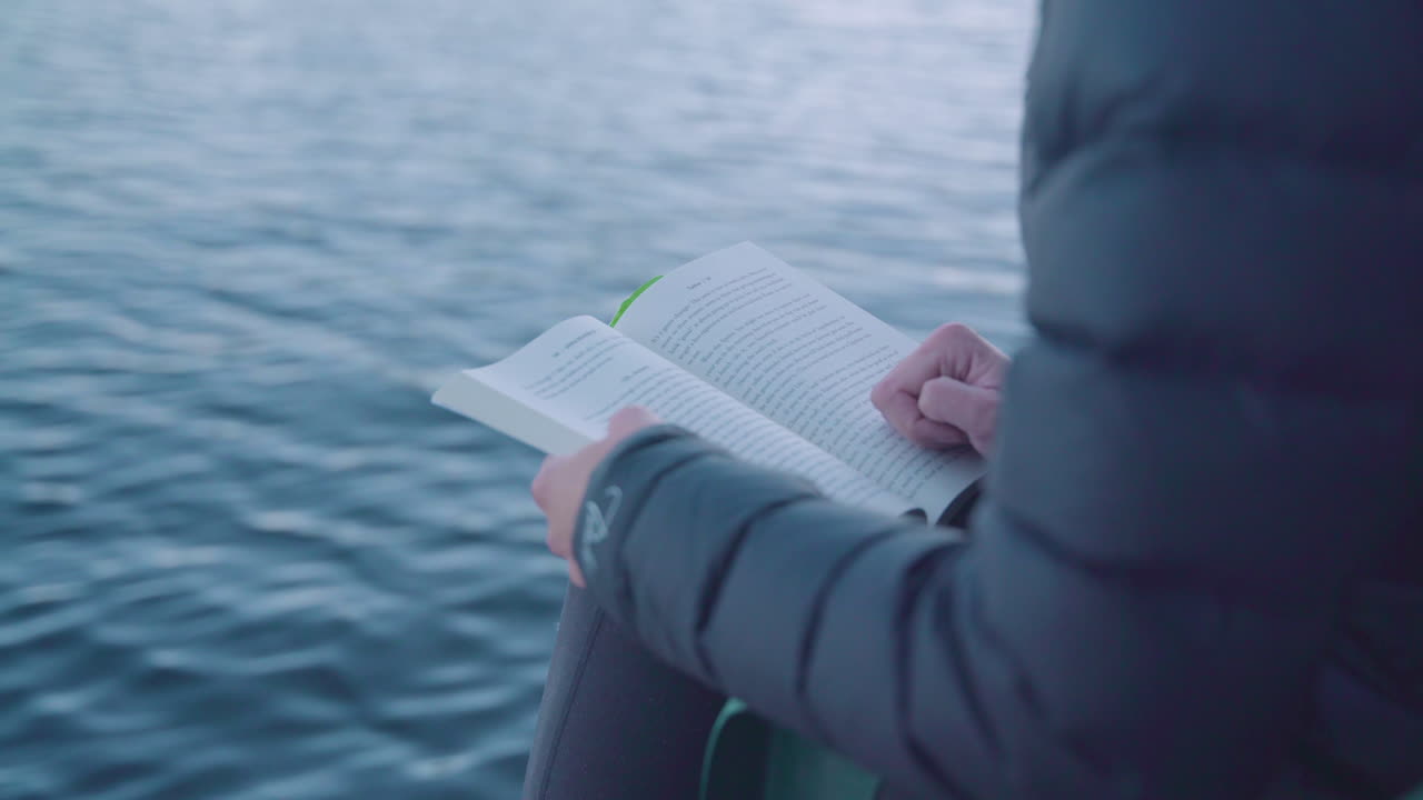 Donna che legge un libro vicino all'acqua con un cappotto invernale