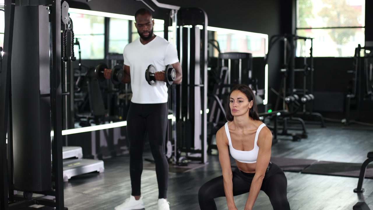 Frau macht Kniebeugen mit Kettlebell, während Mann Kurzhanteln im Fitnessstudio curlt