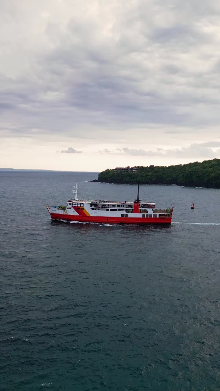 A single ferry leaves Padang Bai Harbor in Bali, sailing across calm turquoise waters toward the open sea. The vertical drone view captures coastal scenery and tropical island travel vibes