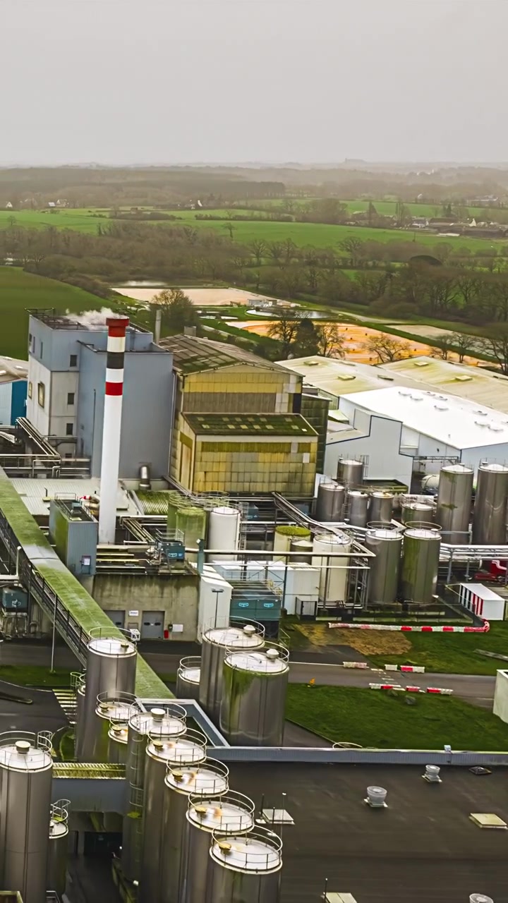 Vertical timelapse footage shows industrial dairy processing plant with storage tanks striped chimney and silos surrounded by green farmland