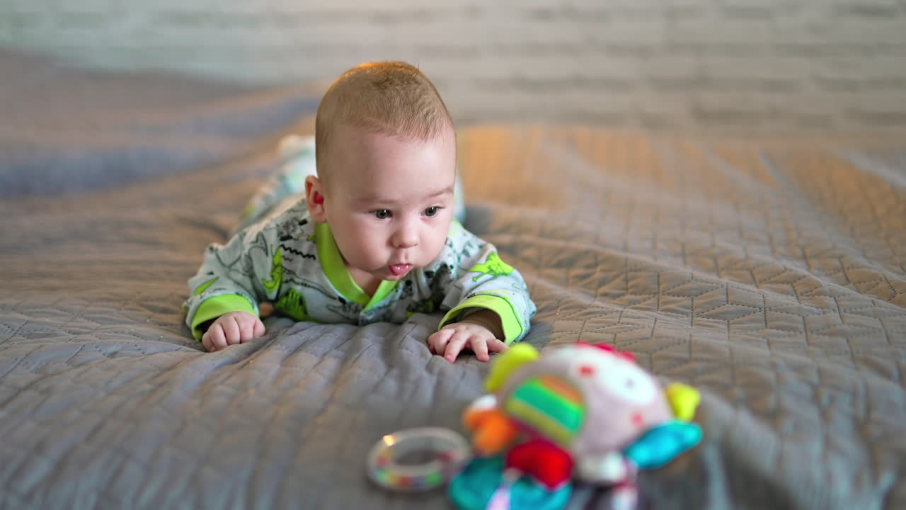 Bright soft toy lies on bed at the foreground. Cute adorable baby boy is interested in a toy and looks at it intently.