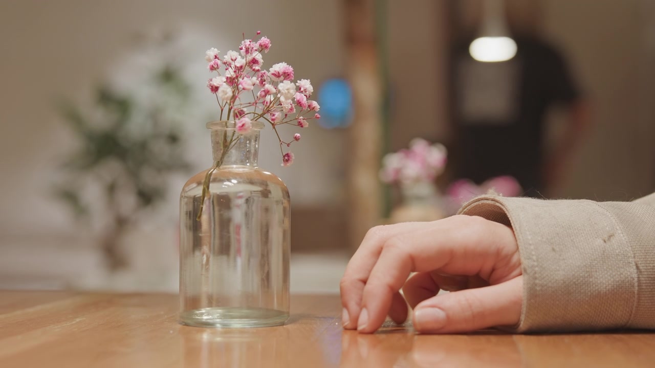 Gros plan sur une main posée sur une table en bois, près d'un vase en verre transparent contenant des fleurs roses, avec un éclairage doux et un arrière-plan flou. La main bouge légèrement, de manière minimale, dans un cadre intérieur serein.
