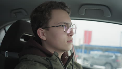 white boy wearing glasses looks out car window as blurred traffic passes by in urban winter setting inside vehicle cabin interior with soft natural light reflective gaze and contemplative mood