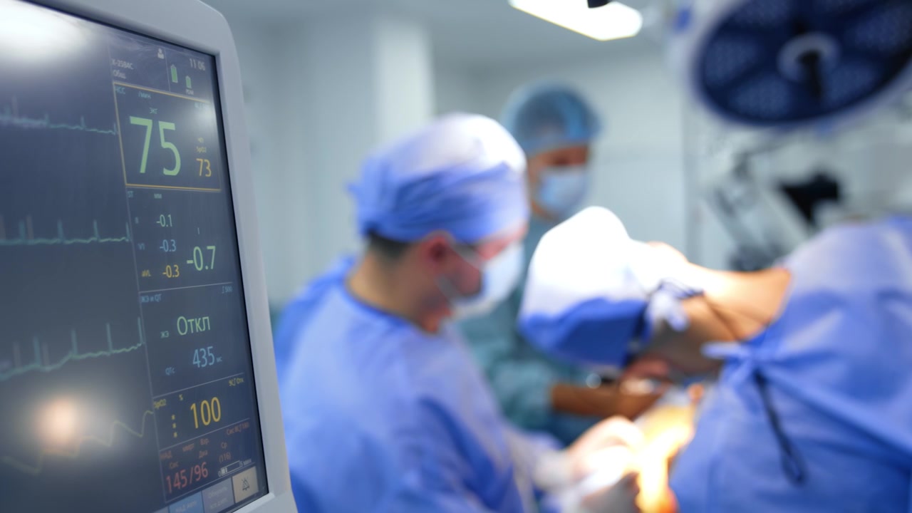 Alcuni numeri sul monitor di un ventilatore polmonare in funzione durante un'operazione. Un team di chirurghi uomini lavora sullo sfondo sfocato.