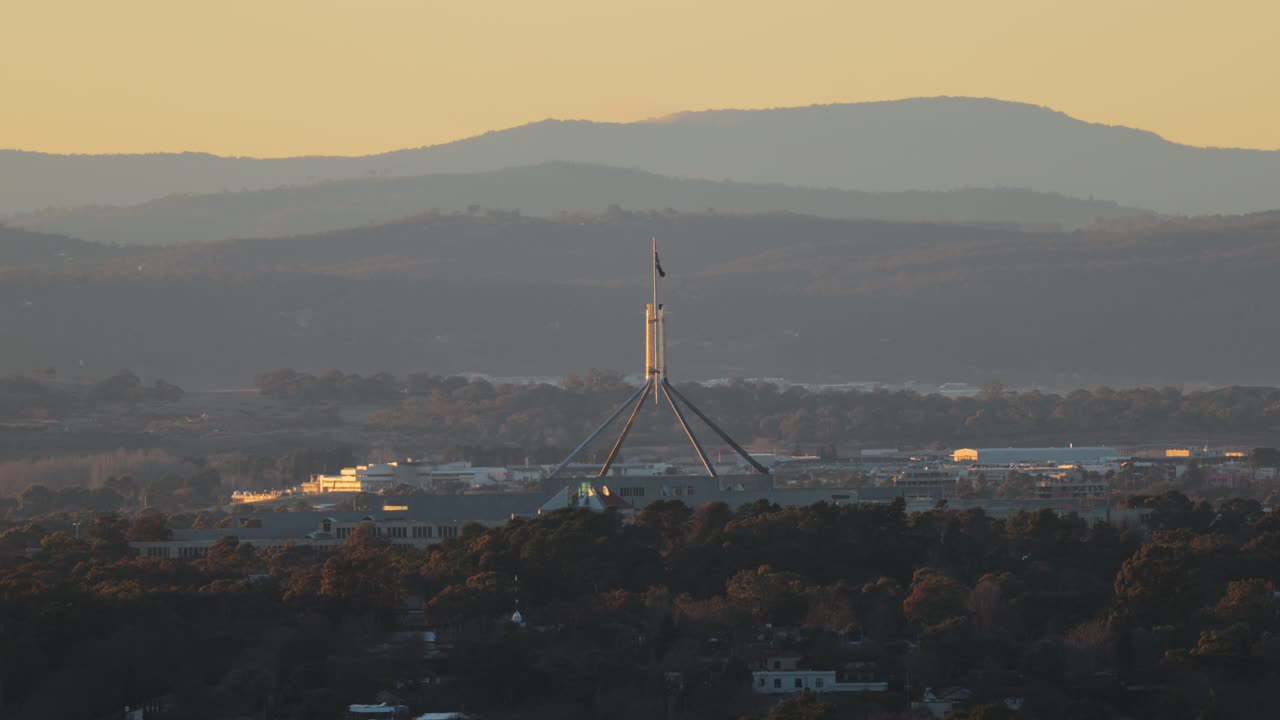 Distant view of Parliament House at dawn with the Australian flag fluttering gently above the building, basking in warm early light