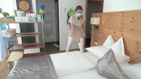 a maid with face mask cleans the hotel room and makes the bed