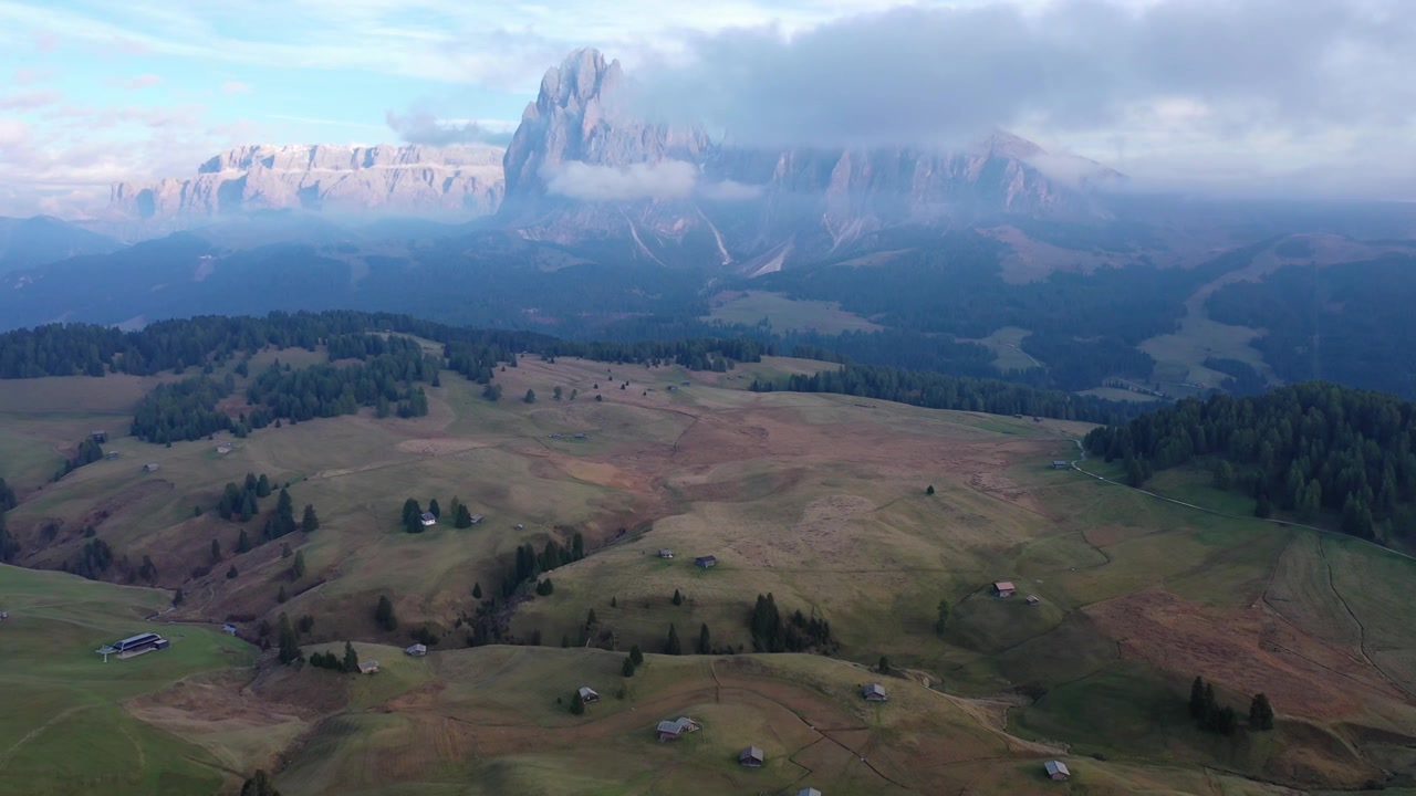 Betagende droneoptagelse fra høj vinkel af de bølgende grønne græsgange og alpehytter ved Alpe di Siusi i de italienske Dolomitter. Med de majestætiske Sassolungo-bjergtoppe under overskyet sommerhimmel.