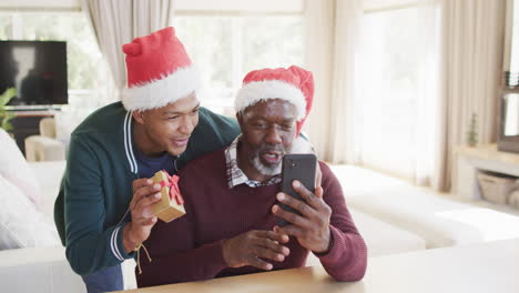 Feliz-Padre-E-Hijo-Afroamericanos-Con-Sombreros-Navideños-Teniendo-Videollamadas-Con-Teléfonos-Inteligentes,-Cámara-Lenta