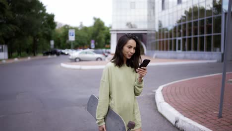 A-brunette-girl-in-a-green-sweater-walks-in-the-park.-The-girl-has-a-skateboard-in-one-hand-and-a-phone-in-the-other.-The-girl