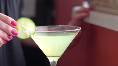 cucumber wheel slice garnish is placed on a martini glass filled with a green cocktail or mocktail drink closeup, alcohol mixed drink made by woman bartender with pink nailpolish hands