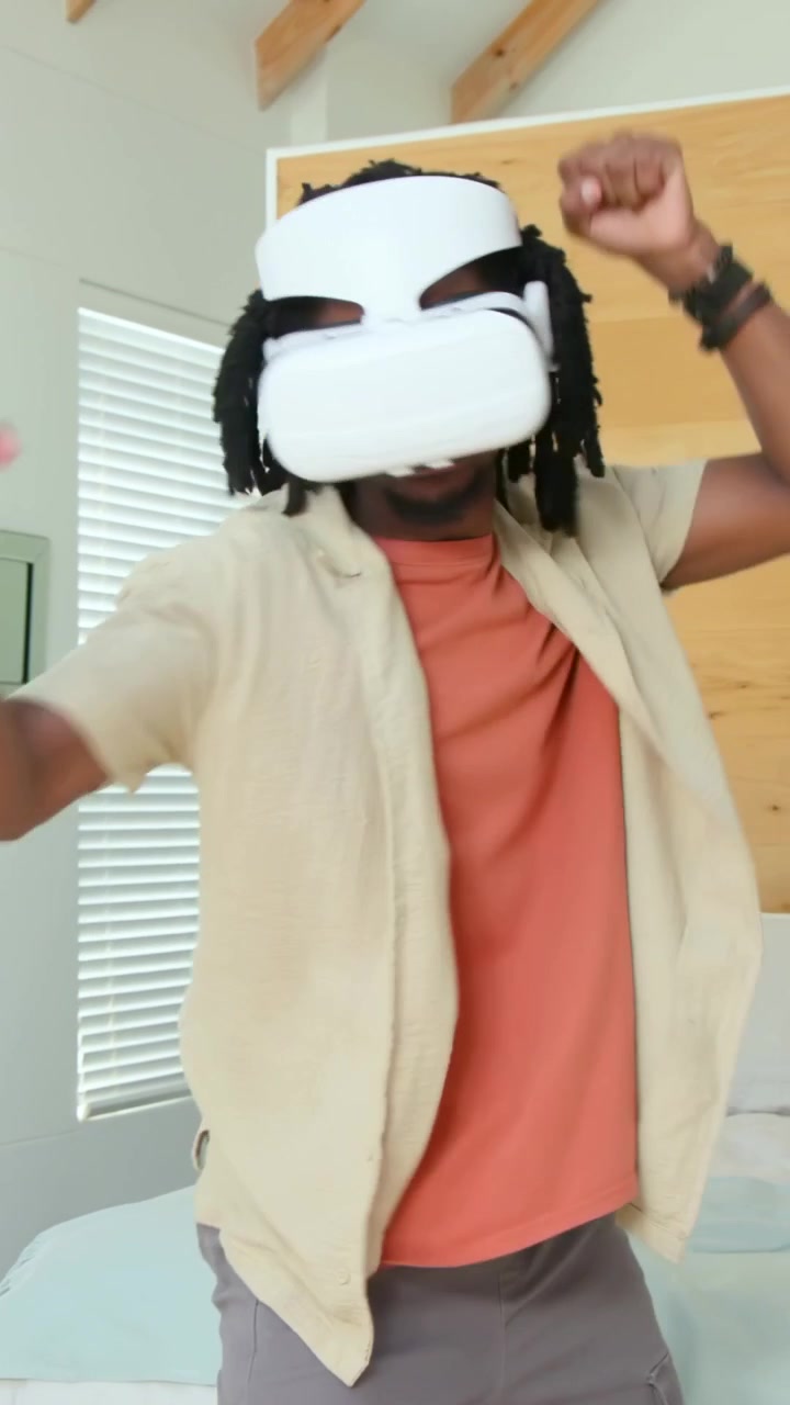 Video verticale: Uomo che reagisce a un segnale VR, indossa una maglietta beige e cuffie bianche, fa boxe e si muove nella camera da letto.
