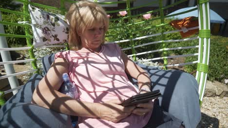 woman sits in comfy swing garden furniture reading digital ebook novel  relaxing on warm sunny day. left to right slide