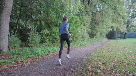 dynamic shot: camera follows active woman running through park on path, grassfield on right, forest on left