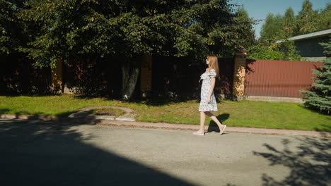 side view of woman in floral dress walking slowly toward tree on residential sidewalk, sun casting shadows of leaves on pavement, calm outdoor setting with brick fence and green lawn