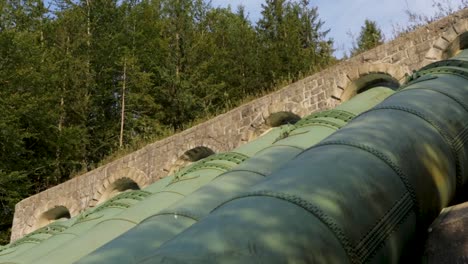 large green pipeline running over a stone bridge in a forested area, sunlight filtering through trees