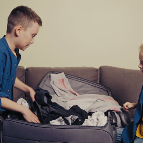 happy brothers packing a big suitcase with clothes at home. two cute boys in blue shirts putting many clothes into the travelling bag while sitting on the sofa in the room. slow motion.