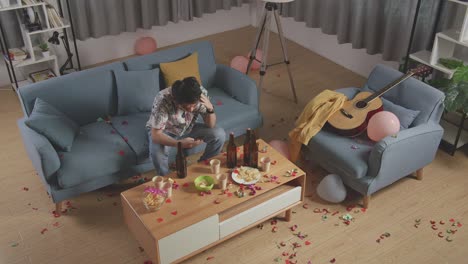 vista superior de un hombre asiático borracho sentado en un sofá y jugando con su teléfono inteligente después de tener una fiesta en casa